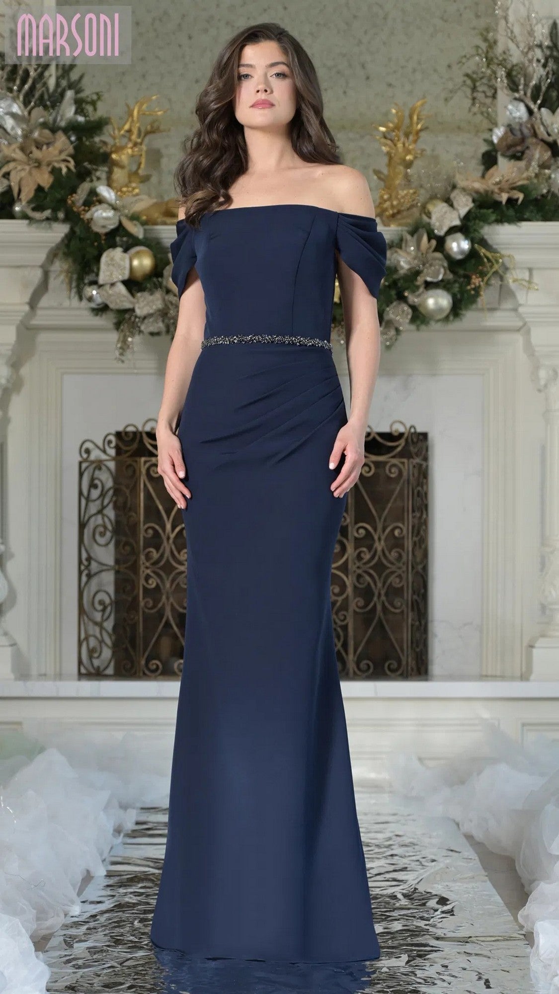 A woman stands indoors wearing the midnight blue Marsoni MV1352, a floor-length formal stretch-crepe off-shoulder gown with a shimmering beaded waistband. Ornate décor and floral arrangements are visible in the background.